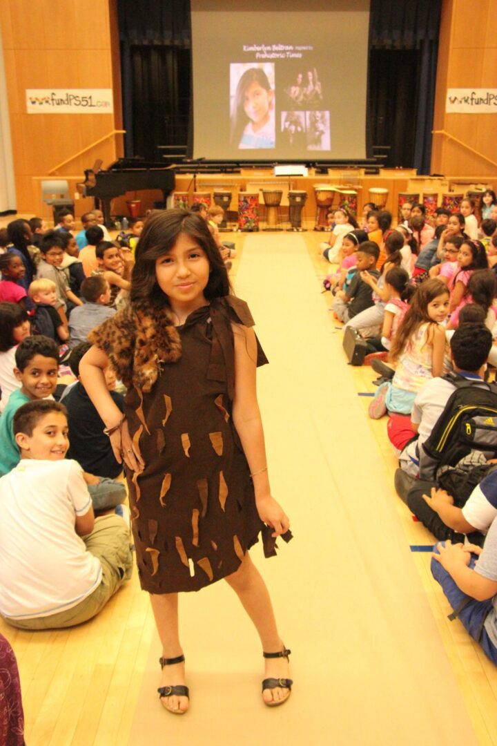 A girl in a brown dress standing in front of a crowd.