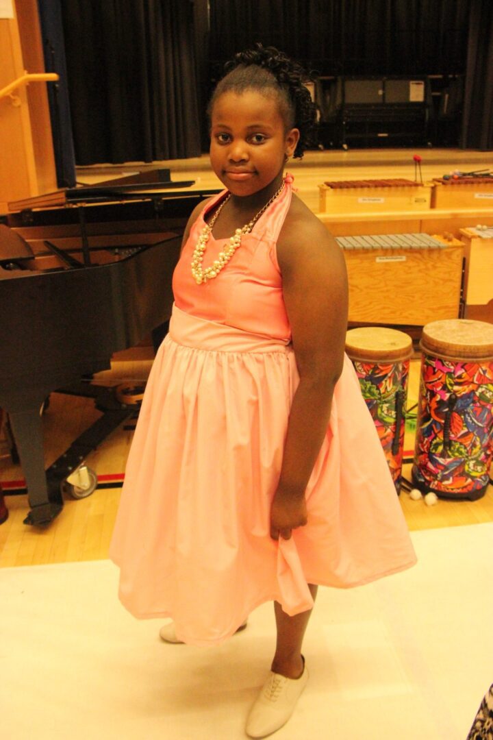 A young girl in a pink dress standing in front of a piano.