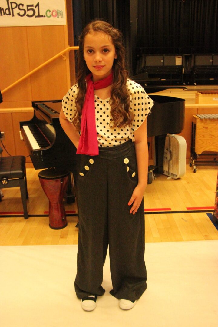 A young girl in polka dot pants standing in front of a piano.