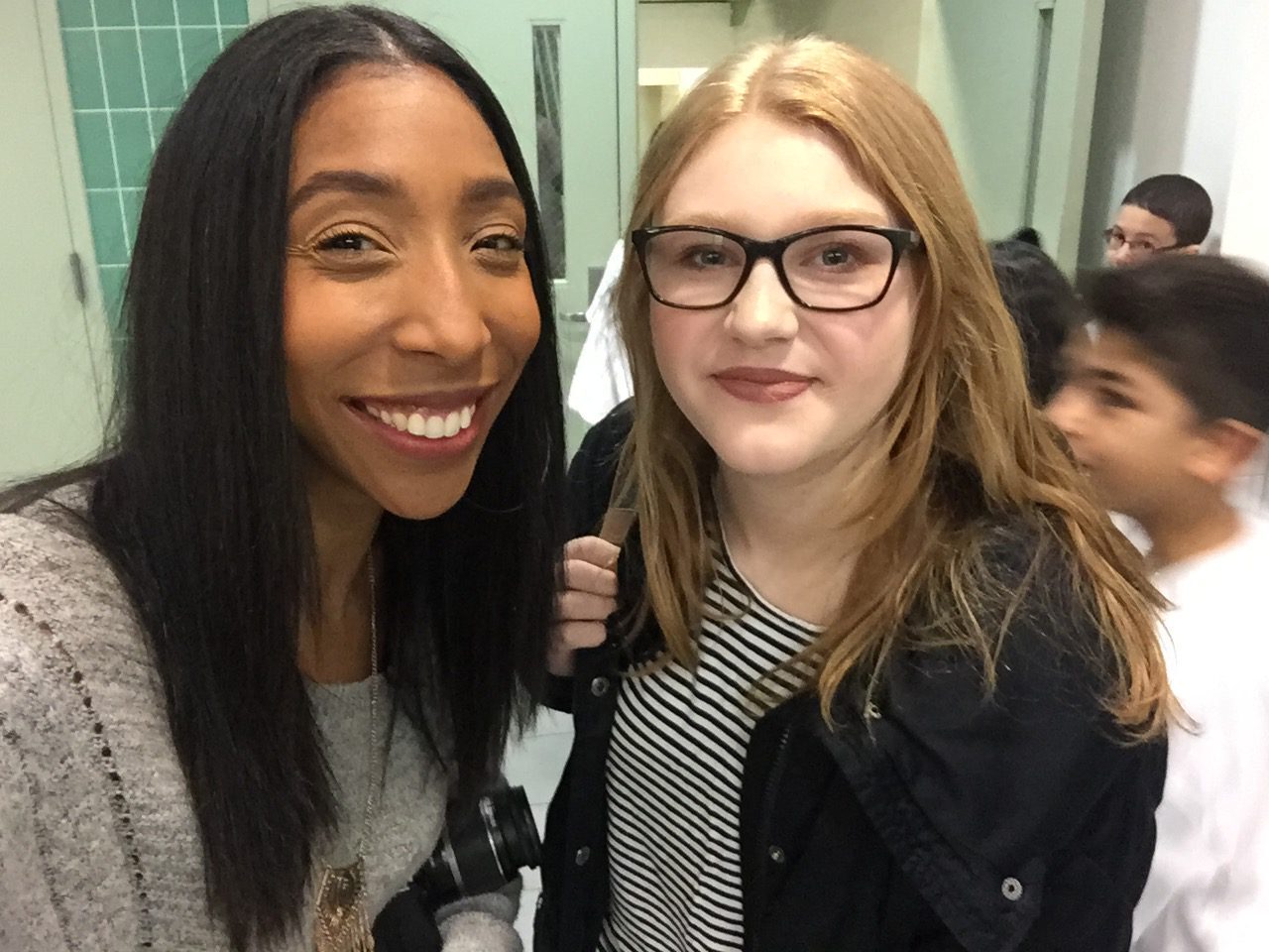 A girl smiles while standing next to another girl in a hallway.