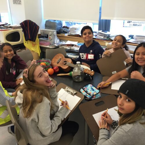 A group of diverse middle school students sitting at a table with art supplies and musical instruments, smiling and interacting in a classroom.
