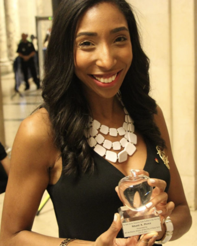 A woman smiling and holding an award at an indoor event, wearing a black sleeveless top and a white necklace.
